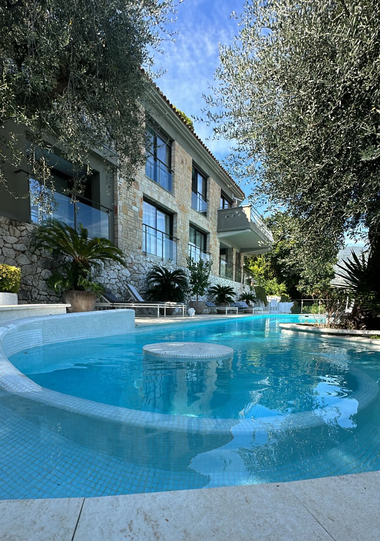 Piscine aux formes courbes en mosaïque claire, au pied d'une villa en pierre